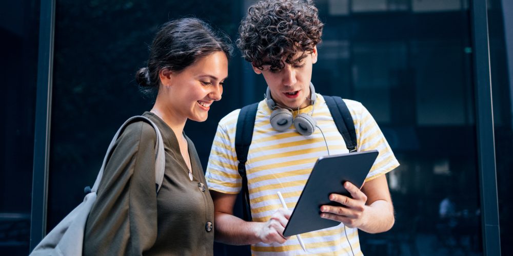 Portrait of a Couple of Students Standing and Using the Digital Tablet A couple of cheerful students outdoors, standing next to each other. Them man is using the digital tablet and drawing something on it.